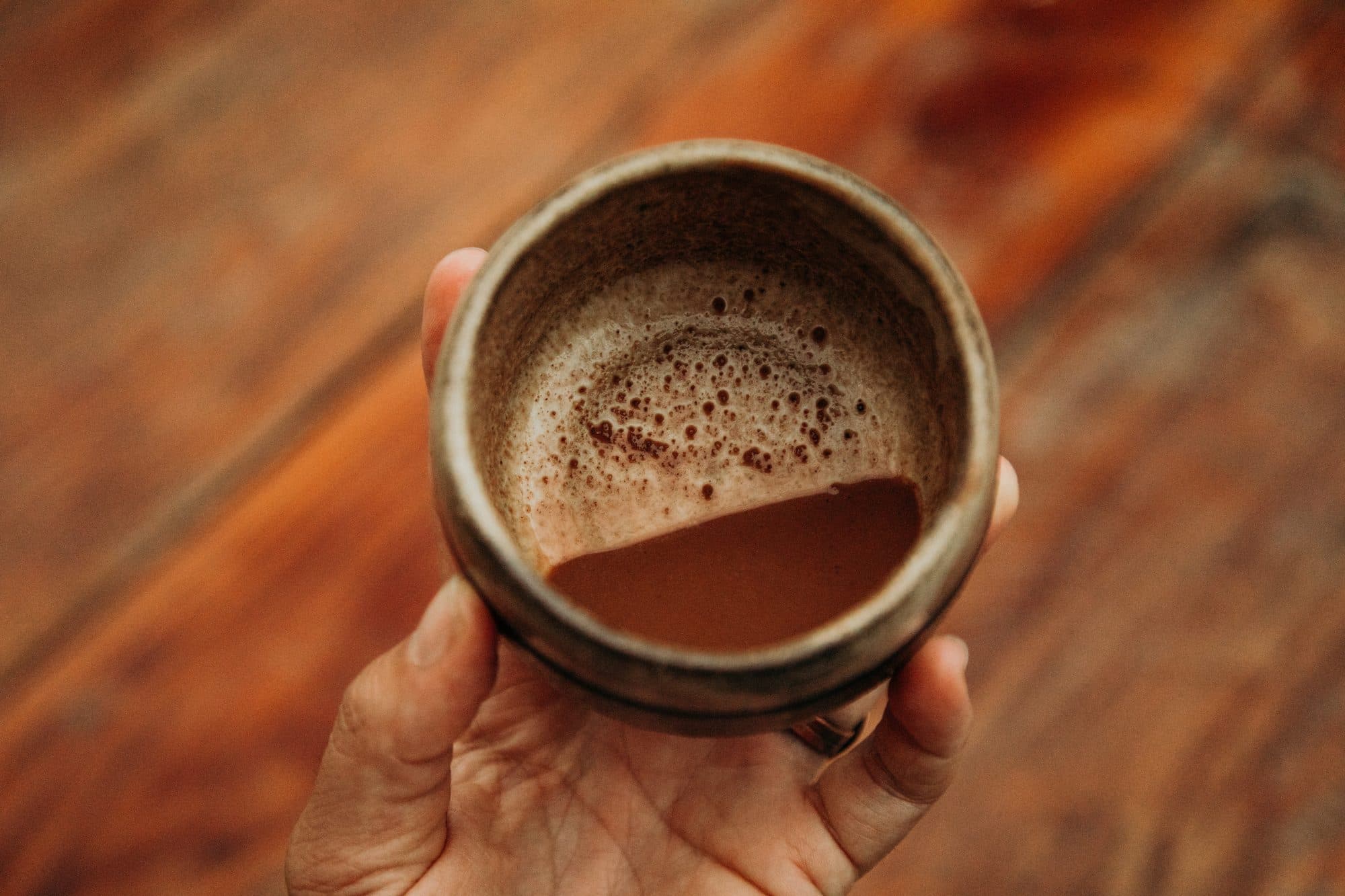 A hand-thrown cup of frothy cacao held over a wooden table